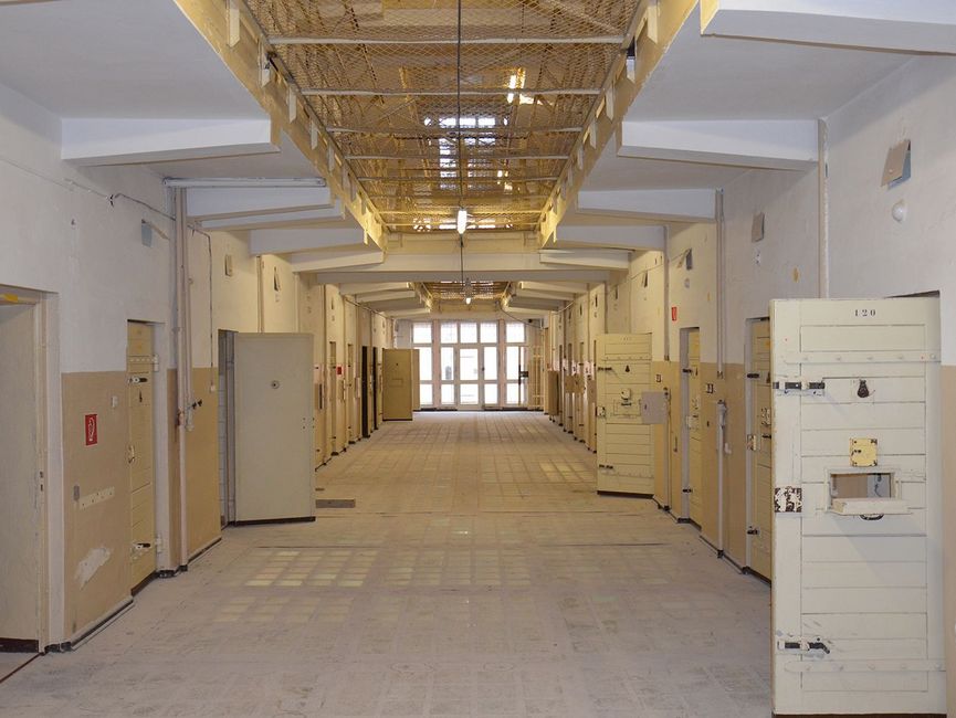 Besucher durchschreiten die Korridore der Haftanstalt. Die Zellentüren links und rechts sind geöffnet.