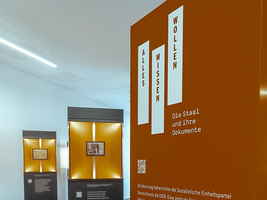 Blick in die Wanderausstellung. Zu sehen sind zwei Stelen mit Glasvitrinen. Im Vordergrund steht eine orangene Tafel mit Informationen über die Ausstellung.