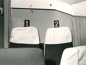 Sitzplätze in einem Flugzeug, die jeweils mit einem Nummernschild markiert sind. In der Wand ist ein Einschussloch zu sehen.