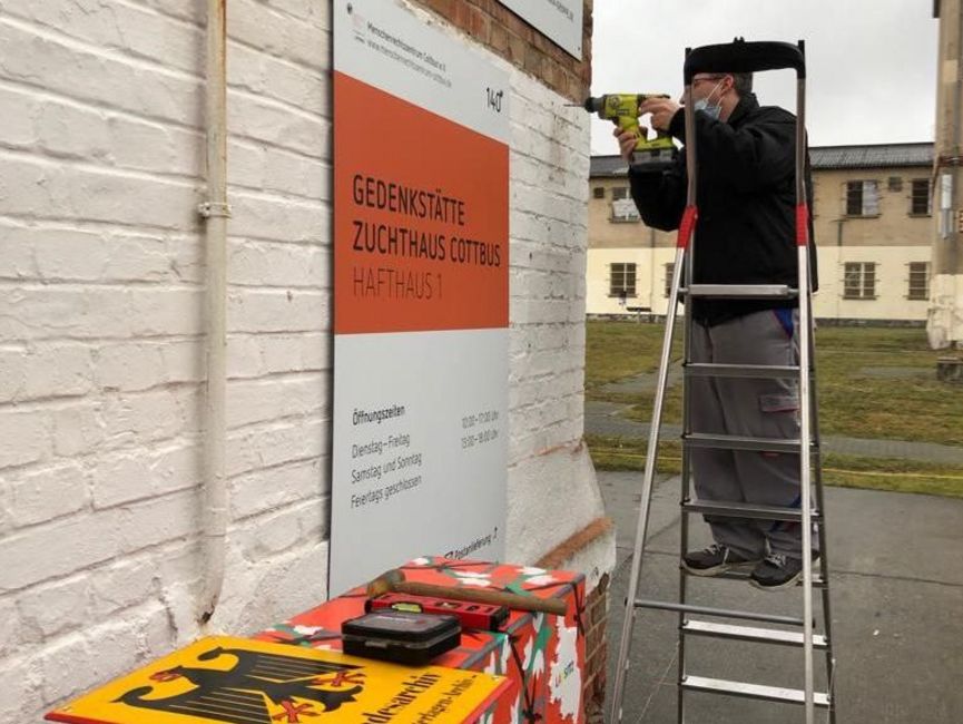  An den Mauern der Gedenkstätte Zuchthaus Cottbus steht ein Mann auf einer Leiter und befestigt ein Schild des Bundesarchivs.