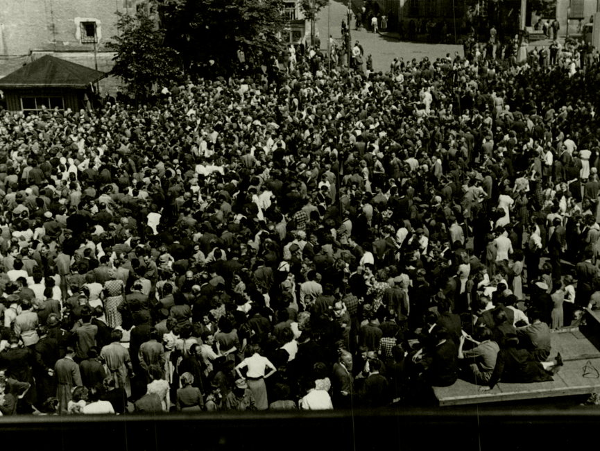 Erneut der Marktplatz im thüringischen Sömmerda am 17. Juni 1953, aufgenommen aus erhöhter Perspektive. Die Menschenmenge erscheint aus diesem Blickwinkel und zum Zeitpunkt der Aufnahme wieder etwas dichter. Im Vordergrund eine der Bretterbuden, auf der einige junge Männer sitzen.