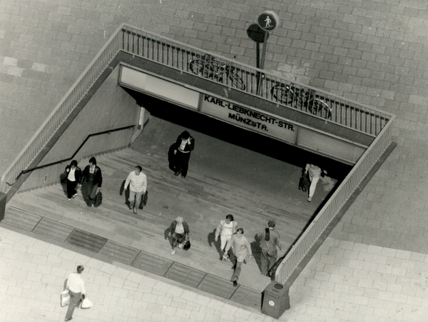 [Auf dem schwarzweißen Lichtbild lassen sich sehr gut einige Menschen erkennen, die die Treppen zur U-Bahnstation hinunter- oder heraufgehen. Auf dem Schild über den Treppen  ist zu lesen "Karl-Liebknecht-Str. Münzstr.".]