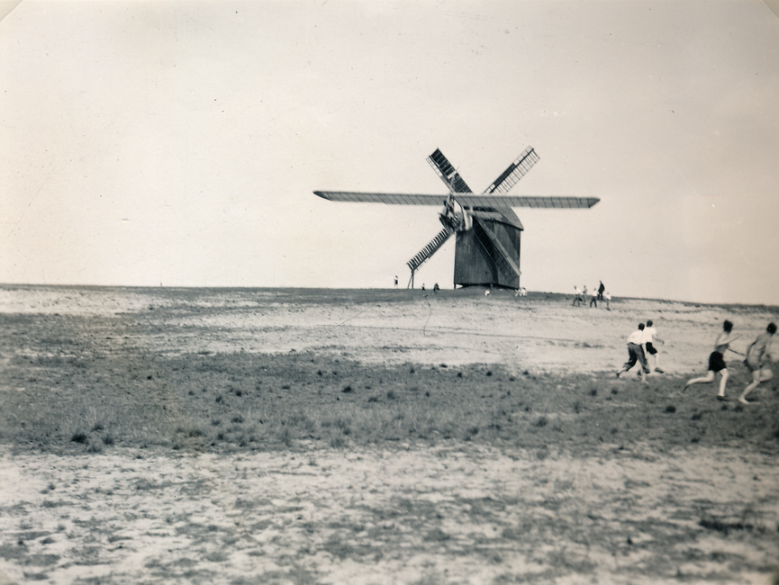 Auf dem schwarz-weißen Lichtbild ist im Hintergrund eine Windmühle zu sehen, die auf freiem Feld steht. Direkt davor befindet sich ein Gleitflieger in der Luft. Des weiteren befinden sich einige Schaulustige im Bild.