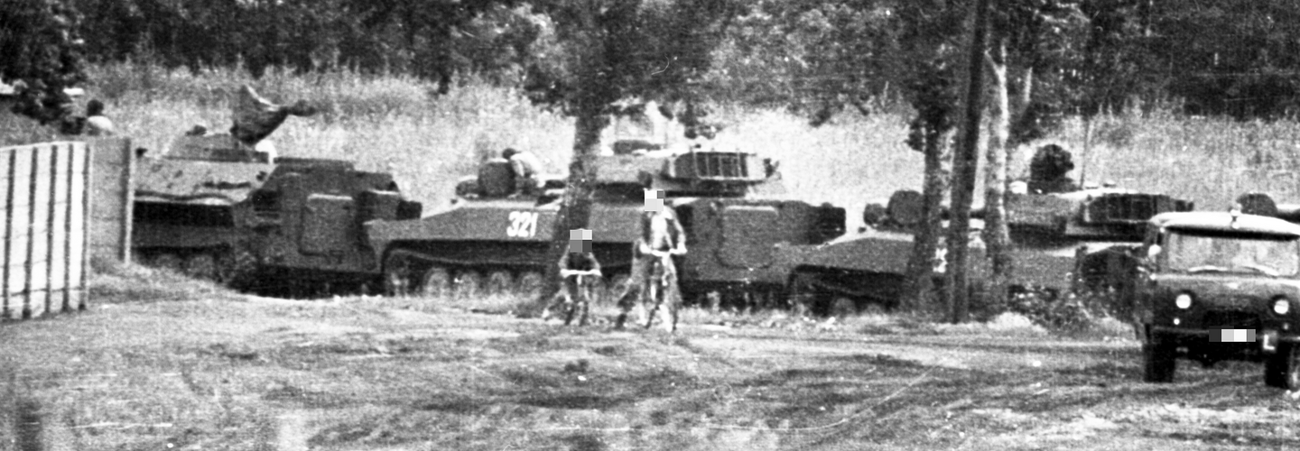 Neben einer Kolonne aus drei Panzern und einem weiteren Militärfahrzeug sind ein Kind und ein Mann auf Fahrrädern zu sehen. Bäume im Hintergrund weisen auf ein bewaldetes Gebiet hin.