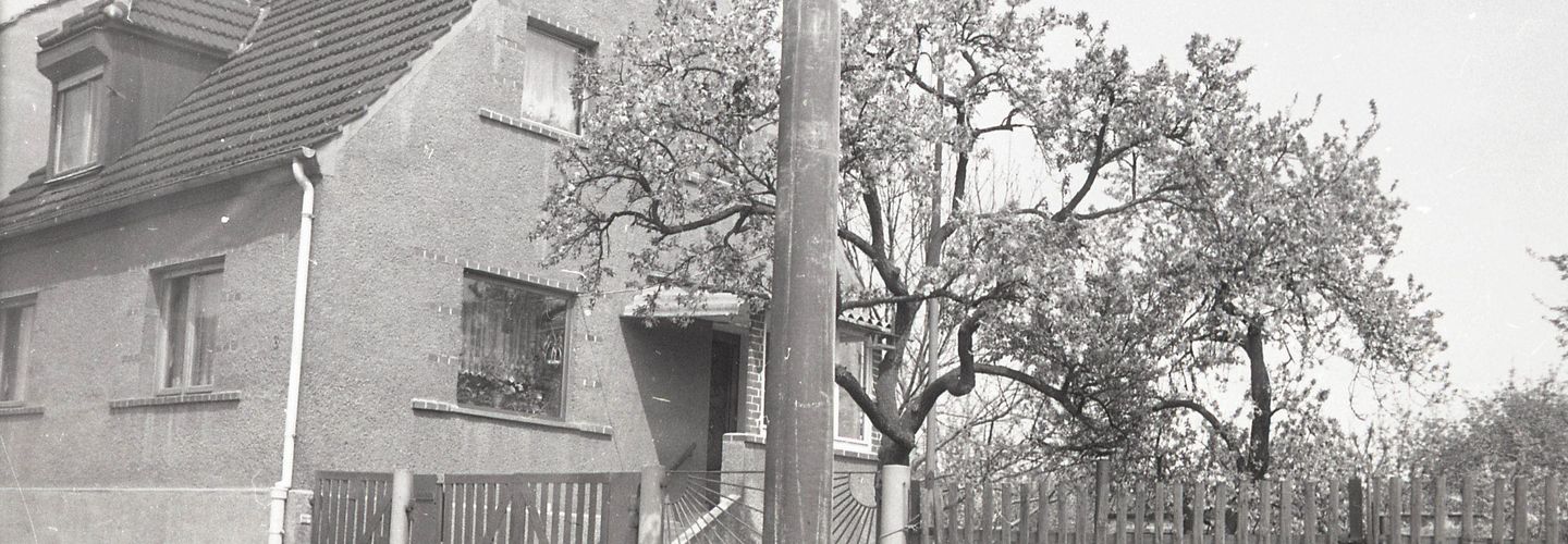 [Die schwarz-weiß Fotografie wurde von der Beifahrerseite eines parkenden Fahrzeuges geschossen. Links auf der Fotografie ist ein Einfamilienhaus an einer Straße zu sehen. Auf dem Grundstück, rechts neben dem Haus, steht ein Kirschbaum. Angrenzend an den Kirschbaum beginnt ein neues Grundstück.]