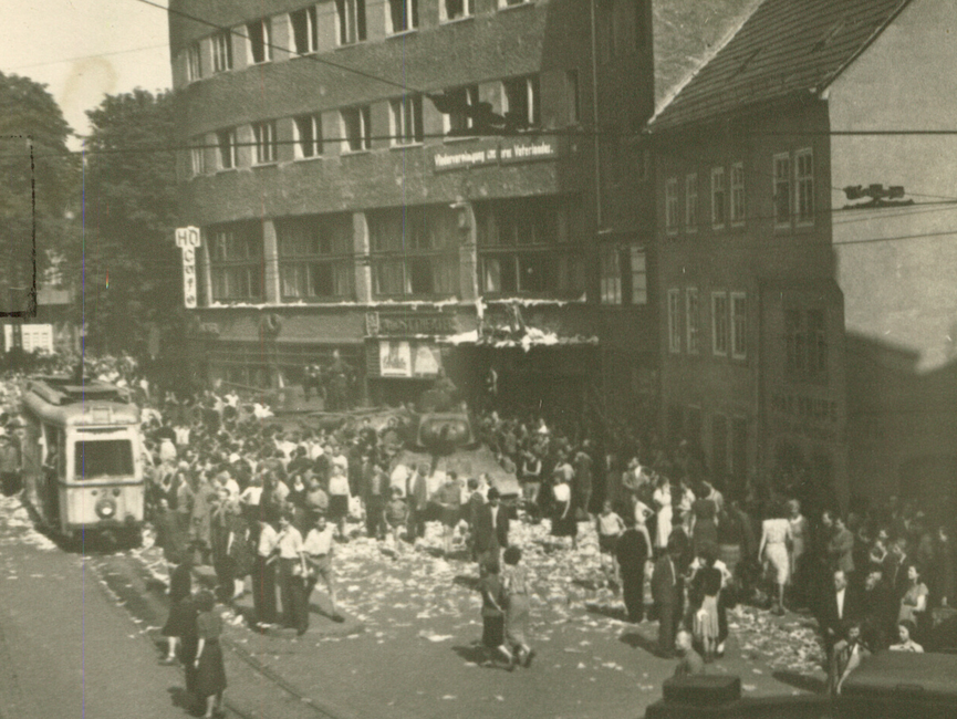 Auf dem Holzmarkt in der Jenaer Innenstadt befinden sich mehrere Menschentrauben, die sich um zwei Panzer ringen. Am linken Bildrand sieht man einen der Straßenbahnwagen, mit denen die Jenaer Bürgerinnen und Bürger den Weg der sowjetischen Panzer blockierten.