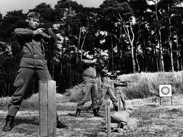 Das schwarzweiße Lichtbild zeigt im Halbprofil vier uniformierte Männer mit Gehörschutz beim Übungsschießen mit automatischen Waffen. Im mittleren Bildgrund steht eine Zielscheibe. Der Übungsplatz ist vor einem Kiefernwald gelegen.