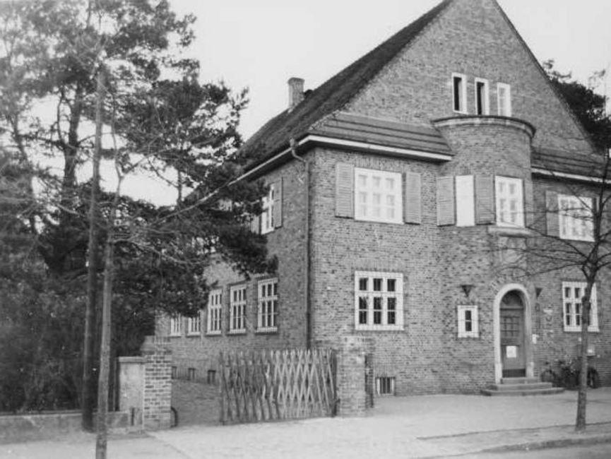 Das schwarz-weiße Lichtbild zeigt ein Backsteingebäude, zwei Etagen mit Spitzdach und Erker mit Eingangstür an der Schmalseite. Hinter einem Jägerzaun links neben dem Haus geht es in eine gepflasterte Auffahrt.