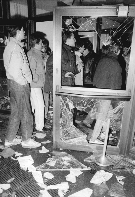 Auf dem Schwarz-Weiß-Bild sind Menschen zu sehen, die durch eine Tür gehen, deren Glas bereits zerschlagen ist. Auf dem Boden liegen Glasscherben und Papier.
