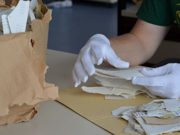 Zwei Hände mit weißen Handschuhen halten Schnipsel von Stasi-Unterlagen in der Hand.