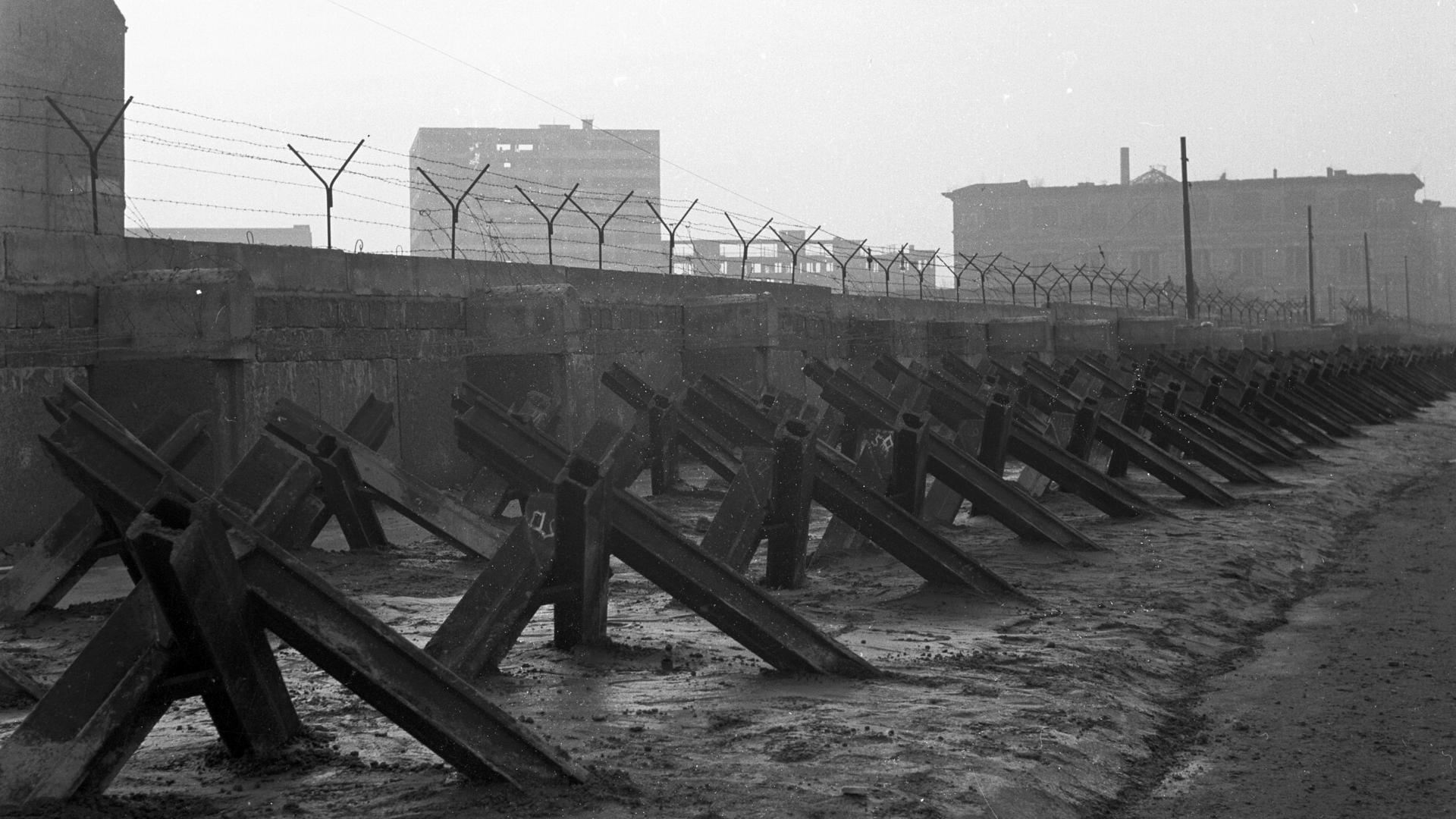 Das Schwarz-Weiß-Foto zeigt den Grenzverlauf in Berlin-Mitte unmittelbar nach dem Mauerbau. Das Bild zeigt eine Mauer mit aufgesetztem Stacheldraht und Panzersperren im Vordergrund. 
