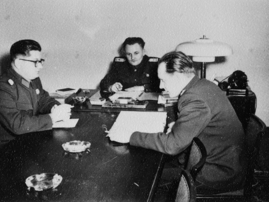 Drei Stasi-Offiziere sitzen an einem Tisch mit Unterlagen.