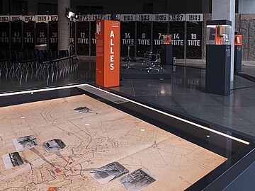 Blick in die Ausstellung "Alles Wissen Wollen". Im Vordergrund ist eine große historische Karte zu sehen. Im Hintergrund stehen mehrere Obejktstelen.