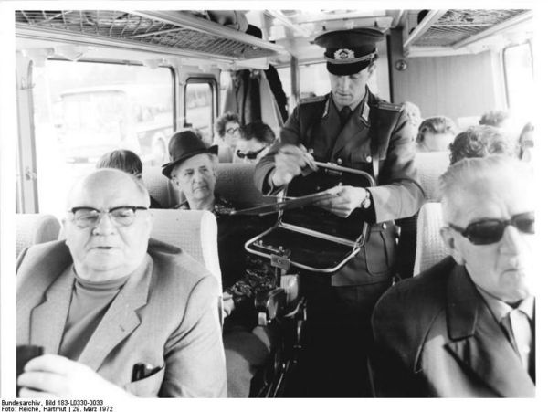 [In einem voll besetzten Bus steht ein Mann in Zolluniform und kontrolliert die Reisedokumente der Passagiere. Um seinen Hals hängt ein Klappfach, er stempelt das Einreisedatum in einen Pass. Es ist ein schwarzweißes Lichtbild.]]