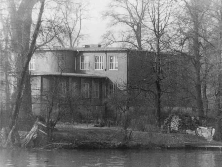 Das schwarz-weiße Lichtbild zeigt ein Gebäude mit Flachdach, das umgeben ist von kahlen Laubbäumen und dicht am Ufer eines Gewässers ist, welches im Bildvordergrund zu sehen ist.
