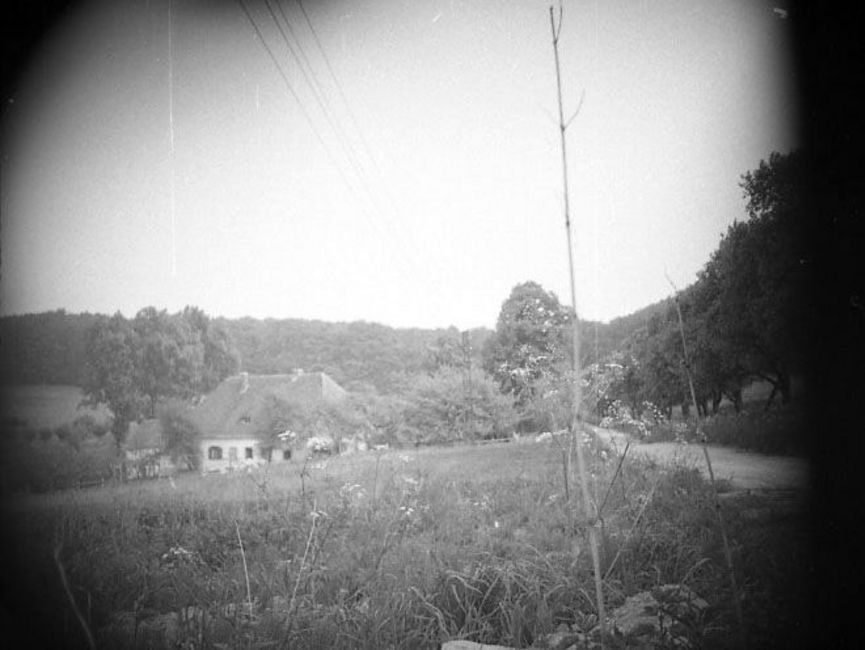 Auf dem schwarz-weißen Lichtbild ist eine hügelige Landschaft abgebildet, im Hintergrund grenzt sie an einen Waldrand. Im Mittelgrund steht ein Haus, umgeben von einigen kleineren Laubbäumen. Die Ecken sind abgedunkelt, vermutlich wurde das Foto verdeckt aufgenommen.