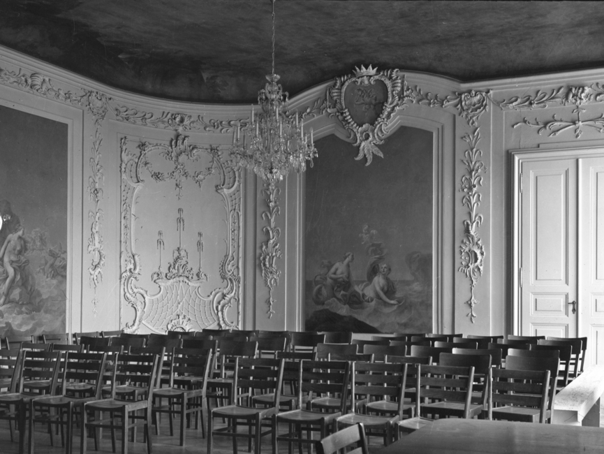 Dieses schwarz-weiße Lichtbild zeigt den "Gartensaal" des Schloss Nischwitz voll bestuhlt. Der Saal im spätbarocken Stil ist mit zwei großen Wandbildern und Stuck verziert.