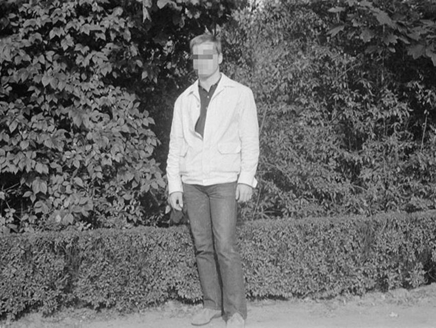 Die Frontalaufnahme zeigt einen Mann in heller Jacke und dunkler Hose vor einer Hecke. Es handelt sich um ein schwarz-weißes Lichtbild.