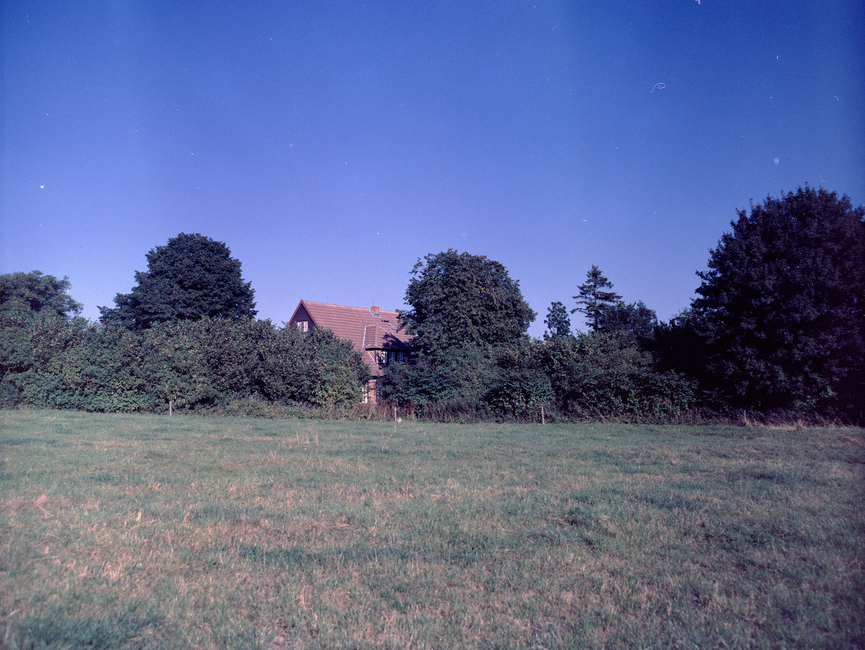 [Die Landschaftsaufnahme zeigt neben viel Wiesenfläche einen sehr blauen Himmel. Das Fachwerkhaus wirkt hinter dichten Bäumen etwas versteckt. Es ist ein Farbfoto.] 