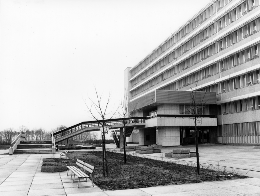 Das Schwarz-Weiß-Bild zeigt einen Teil des Stasi-Krankenhauses in Berlin-Buch. Zu sehen ist der Haupteingang mit einem kleinen Anbau, zu welchem eine Treppe hinaufführt. Im Vordergrund des Bildes ist zudem eine kleine Rasenfläche mit drei kahlen, noch recht kleinen Bäumen zu sehen. Davor steht eine Sitzbank.