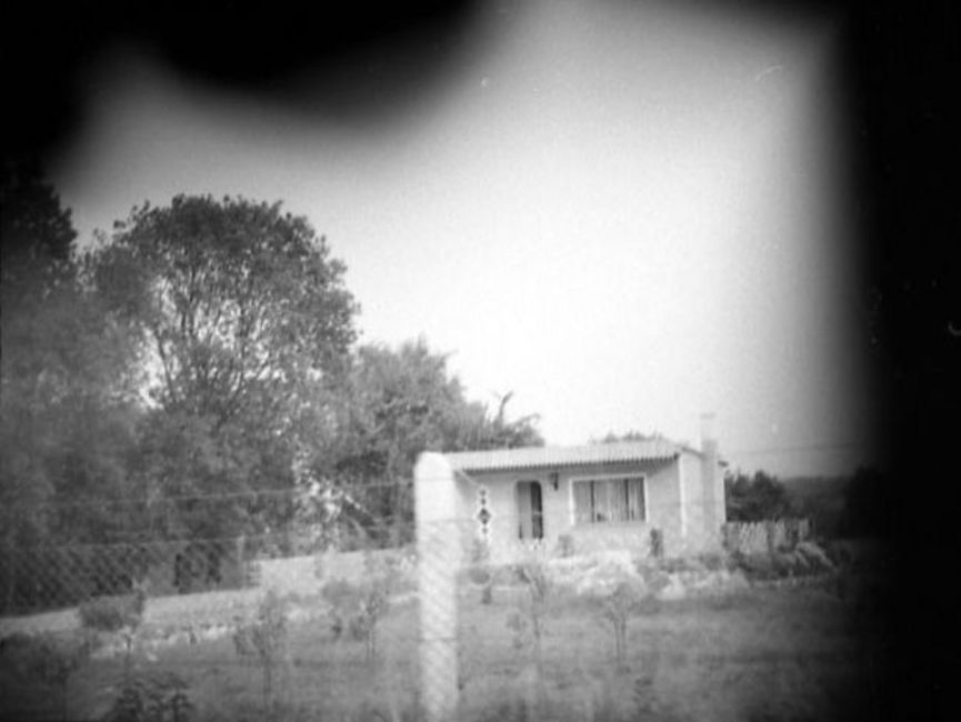 Auf dem schwarz-weißen Lichtfoto wurde ein Bungalow auf einer Anhöhe abgebildet. Das Gelände wird durch einen Maschendrahtzaun abgegrenzt. Die Ecken sind abgedunkelt, vermutlich wurde das Foto verdeckt aufgenommen.