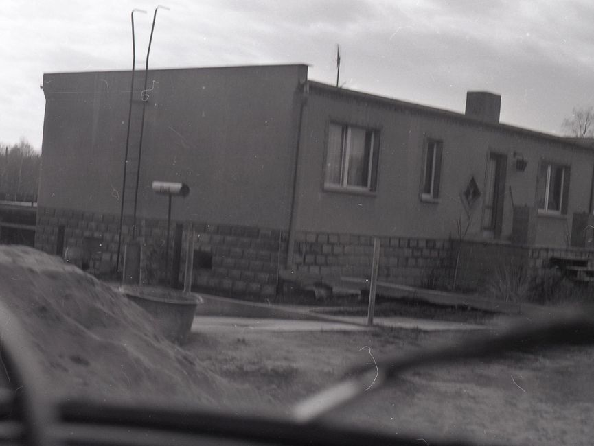 [Die Fotografie ist schwarz-weiß. Durch die Windschutzscheibe eines Fahrzeuges wurde ein einstöckiges Haus fotografiert. Das Haus besitzt an der linken Seite eine Leiter, die auf das Flachdach führt. Hinter dem Haus sind Bäume zu erkennen. Bei dem Haus könnte es sich um das rechte Gebäude von Bild 25 handeln. Im Vordergrund sind unscharf ein Sandhaufen und ein Maurertrog zu erkennen.]