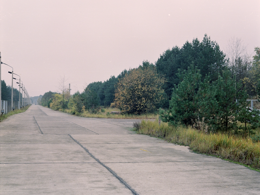 Das bild zeigt eine aus Betonplatten errichtete Straße. Links davon befindet sich ein Zaun mit aufgereihten Straßenlampen. Rechts davon befindet sich Vegetation.