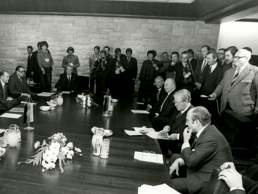 Die Schwarzweißaufnahme zeigt Helmut Schmidt bei der Arbeitsbesprechung der Regierungsdelegationen von BRD und DDR. Hierzu haben sich um einen Besprechungstisch Männer und eine Frau, teils um den Tisch stehend und einige am Tisch sitzend, versammelt. Am rechten Bildrand ist eine zweite Stuhlreihe zu erkennen, die von einigen Männern genutzt wird. Helmut Schmidt blättert gerade in seinen Unterlagen und schaut darauf. Ein Mann hat seine Brille auf sein Schriftstück abgelegt.