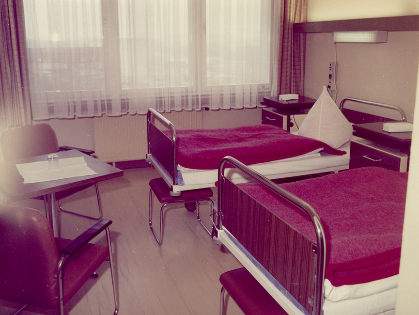 Das Bild zeigt ein Zwei-Bett-Zimmer im Stasi-Krankenhaus Berlin-Buch. Zu sehen sind zwei Betten, auf denen jeweils eine rote, zusammengefaltete Decke liegt. Im Zimmer befindet sich zudem ein Tisch mit zwei Stühlen, zwei Nachttische und Beleuchtung.