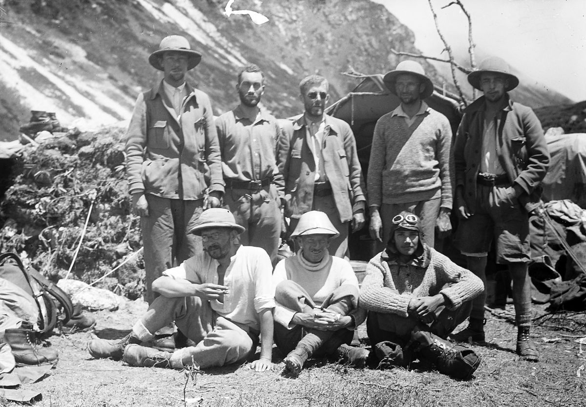 Das Bild zeigt mehrere Männer bei einer Bergexpedition, die für ein Gruppenfoto posieren. Um sie herum sind einige Ausrüstungsgegenstände zu sehen.