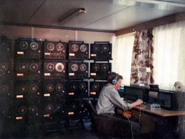 Ein Stasi-Mitarbeiter der Funkaufklärung sitzt mit Kopfhörern an einem Schreibtisch, hinter ihm füllen zahlreiche Aufnahmegeräten die Wand aus.  
