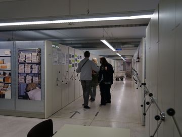 Zwei Besucherinnen und ein Besucher laufen durch das Magazin des Stasi-Unterlagen-Archivs Halle. Links und rechts sind Aktenschränke zu sehen.