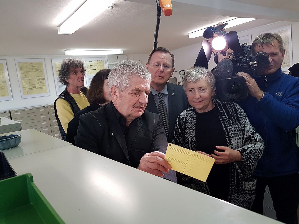 BStU Roland Jahn zeigt Gabriele Stötzer eine Karteikarte. Hinter den beiden steht Ministerpräsident Bodo Ramelow eine Karteikarte. Dahinter befinden sich Journalisten mit Mikrofonen und Kameras.