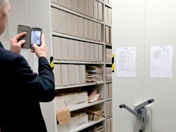 Das Bild zeigt einen Magazinraum mit eingelagerten Unterlagen. Ein Mann fotografiert die Bestände mit seinem Smartphone.