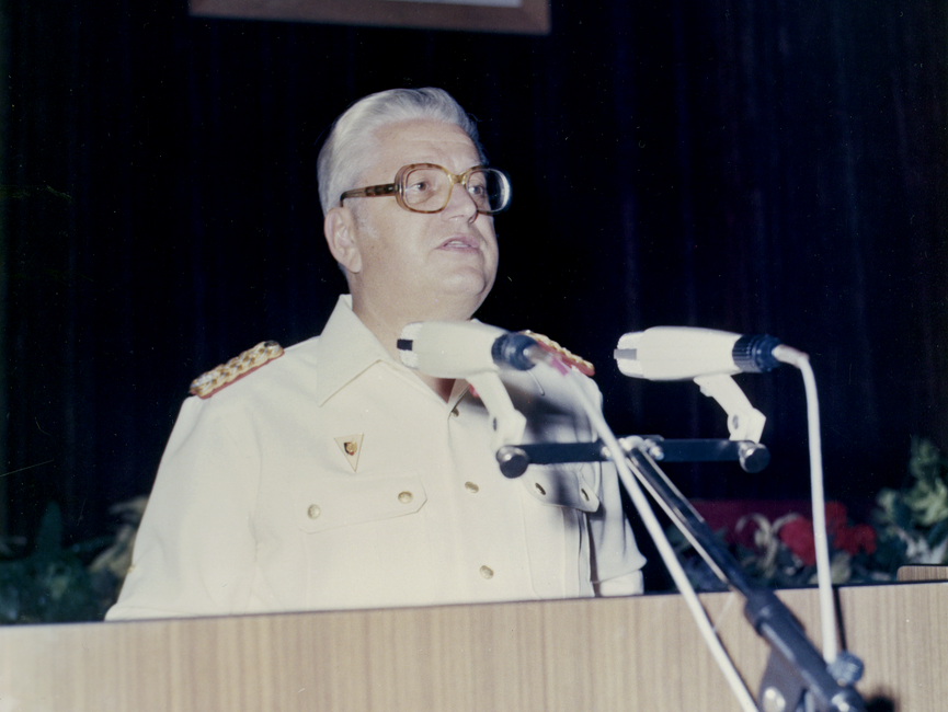 Willi Pösel in weißer Uniform an einem Rednerpult mit Mikrofonen.