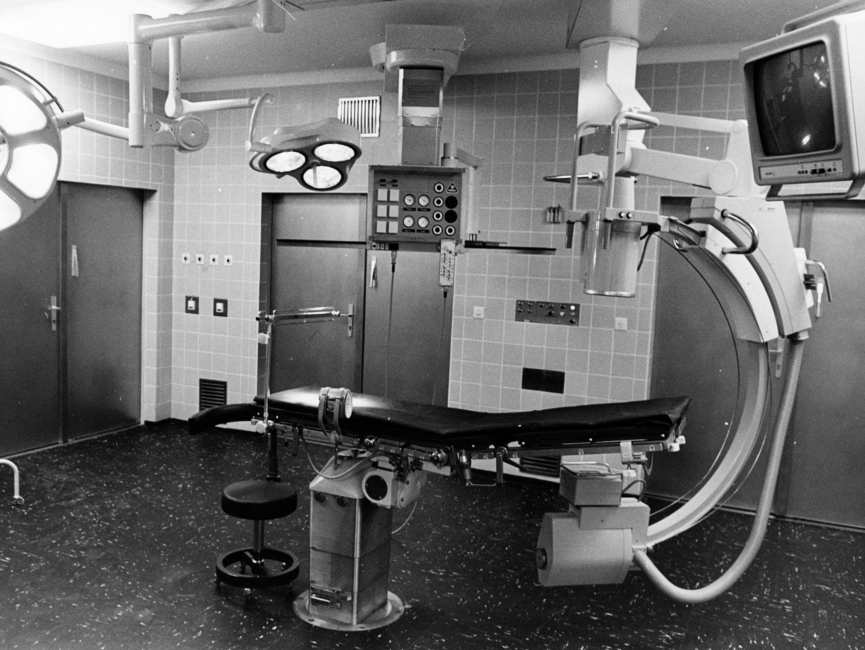 Das Schwarz-Weiß-Bild zeigt einen Operationssaal im Stasi-Krankenhaus Berlin-Buch. In der Mitte des raumes befindet sich eine Liege, rechts daneben ein großes technisches gerät, wahrscheinlich ein Computertomograph. Außerdem befinden sich zwei große Leuchten im Raum.
