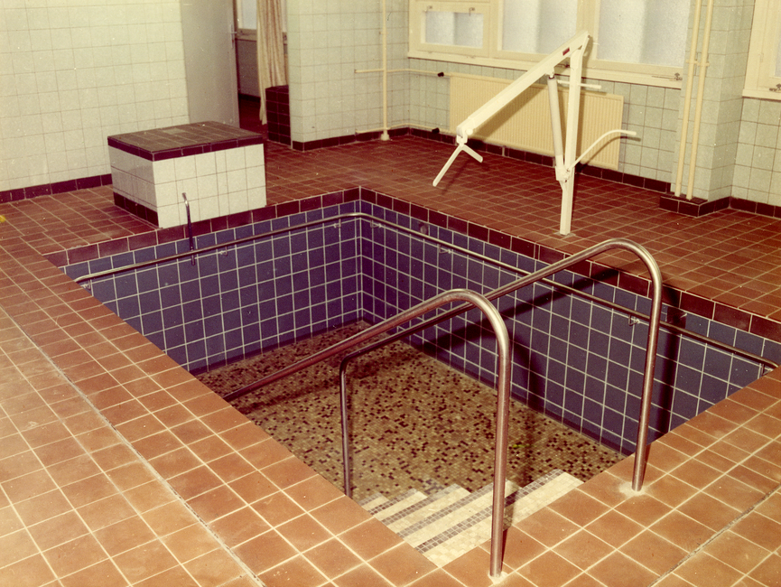 Das Bild zeigt ein kleines Tauch- bzw. Schwimmbecken im Stasi-Krankenhaus in Berlin-Buch. Das Becken befindet sich inmitten eines gefliesten Bereichs. Neben dem Becken befindet sich ein fest installierter Greifarm, bestimmt dazu einen Menschen über Wasser zu halten, um ihm beim Schwimmen zu unterstützen.