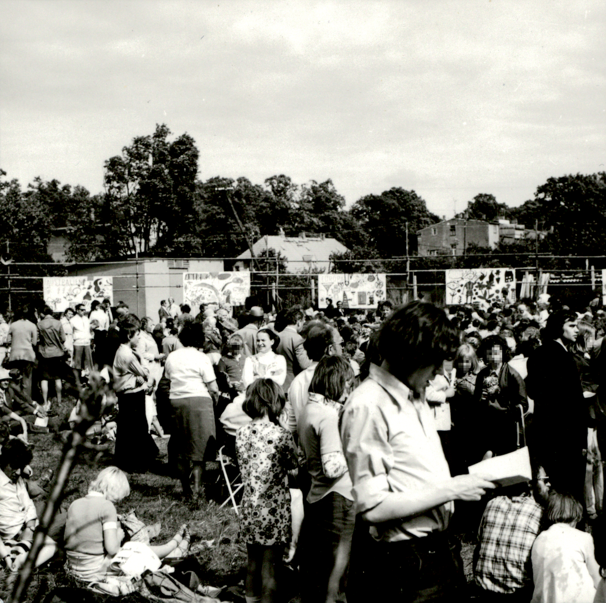 [Das schwarz-weiße Lichtbild zeigt eine Ansammlung von Menschen auf einem freien Platz. Überwiegend stehen die Personen allen Alters auf der Rasenfläche. Im Hintergrund sind kindlich bemalte Transparente an Zäunen angebracht.]