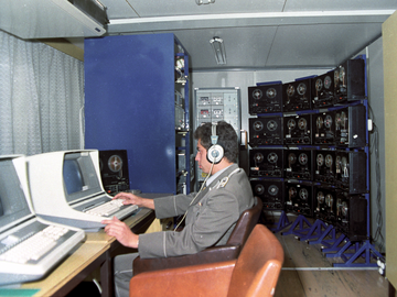 [Das farbige Lichtbild zeigt einen Mann in Uniform im Profil, wie er mit Kopfhörern auf einen schwarzen Computermonitor blickt. Er sitzt in einem Bürosessel am Schreibtisch, neben dem Tischcomputer steht ein Tonbandgerät. Direkt daneben, im Bildvordergrund, ist ein identisch aufgebauter Arbeitsplatz. Im Hintergrund stehen vier Metallständer, bestückt mit jeweils vier Tonbandgeräten. Daneben steht ein Turm mit 14 Kasettenrekordern.]